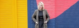 Photo of Victoria Clapham standing against a brightly coloured wall. She has her hands in her coat pockets and is smiling.