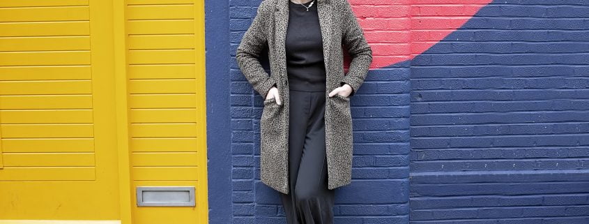 Photo of Victoria Clapham standing against a brightly coloured wall. She has her hands in her coat pockets and is smiling.